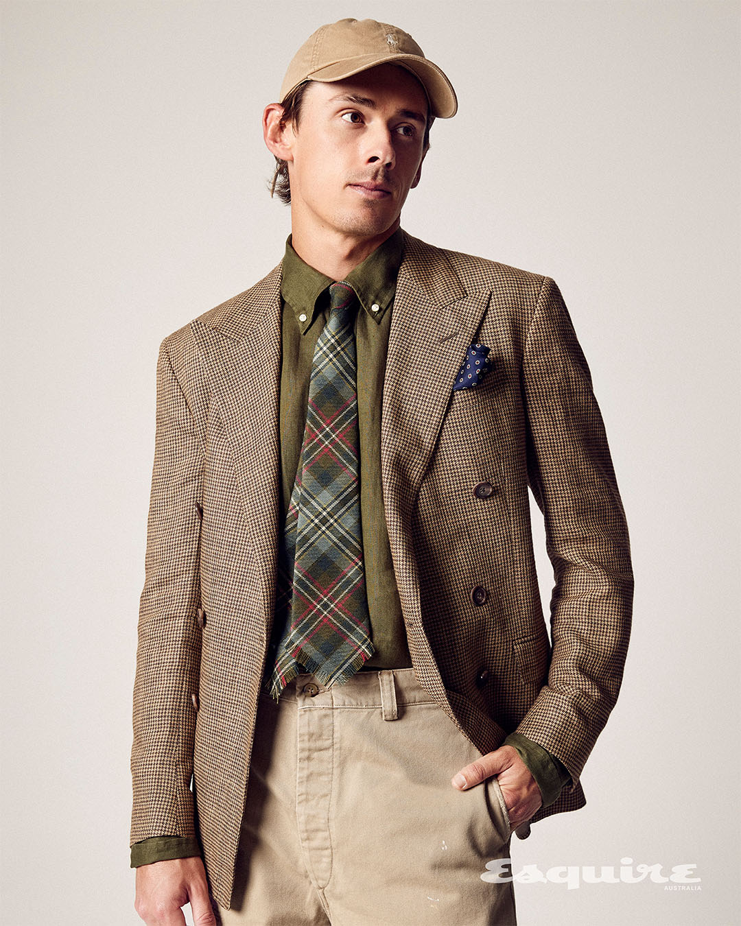 Man in a tan baseball cap, brown plaid blazer, green shirt, and plaid tie posing in a studio with neutral background and soft lighting.