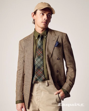 Man in a tan baseball cap, brown plaid blazer, green shirt, and plaid tie posing in a studio with neutral background and soft lighting.