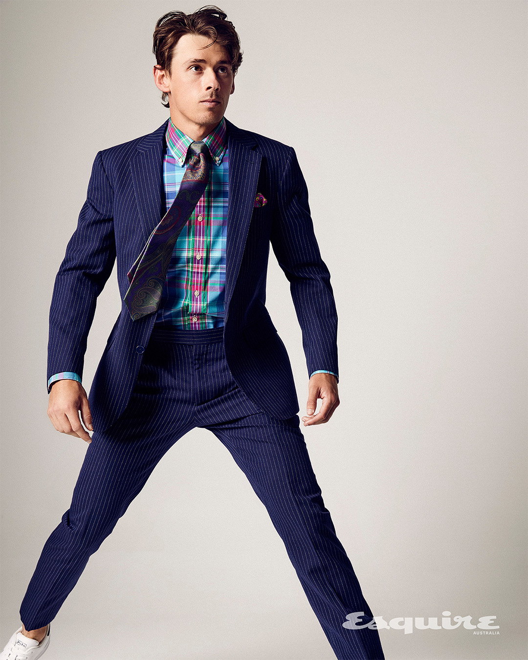 A man in a navy pinstripe suit and white sneakers jumping in front of a light gray backdrop, wearing a colorful plaid dress shirt and tie.