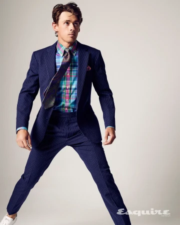 A man in a navy pinstripe suit and white sneakers jumping in front of a light gray backdrop, wearing a colorful plaid dress shirt and tie.