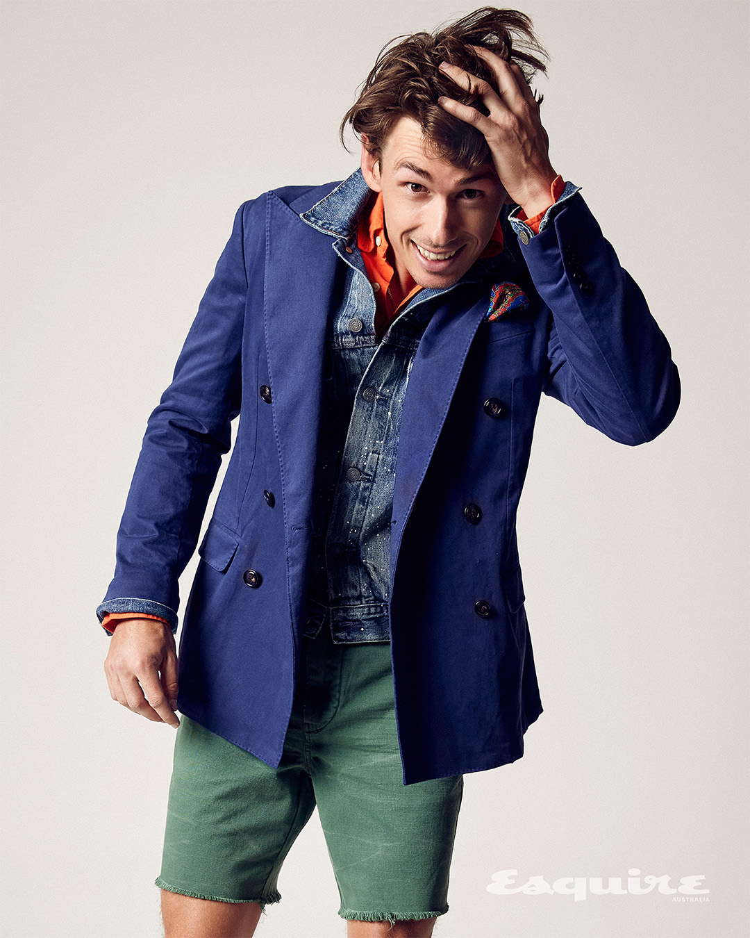 Smiling man in a blue blazer, denim jacket, and green shorts strikes a pose against a neutral backdrop, showcasing a casual yet stylish look.