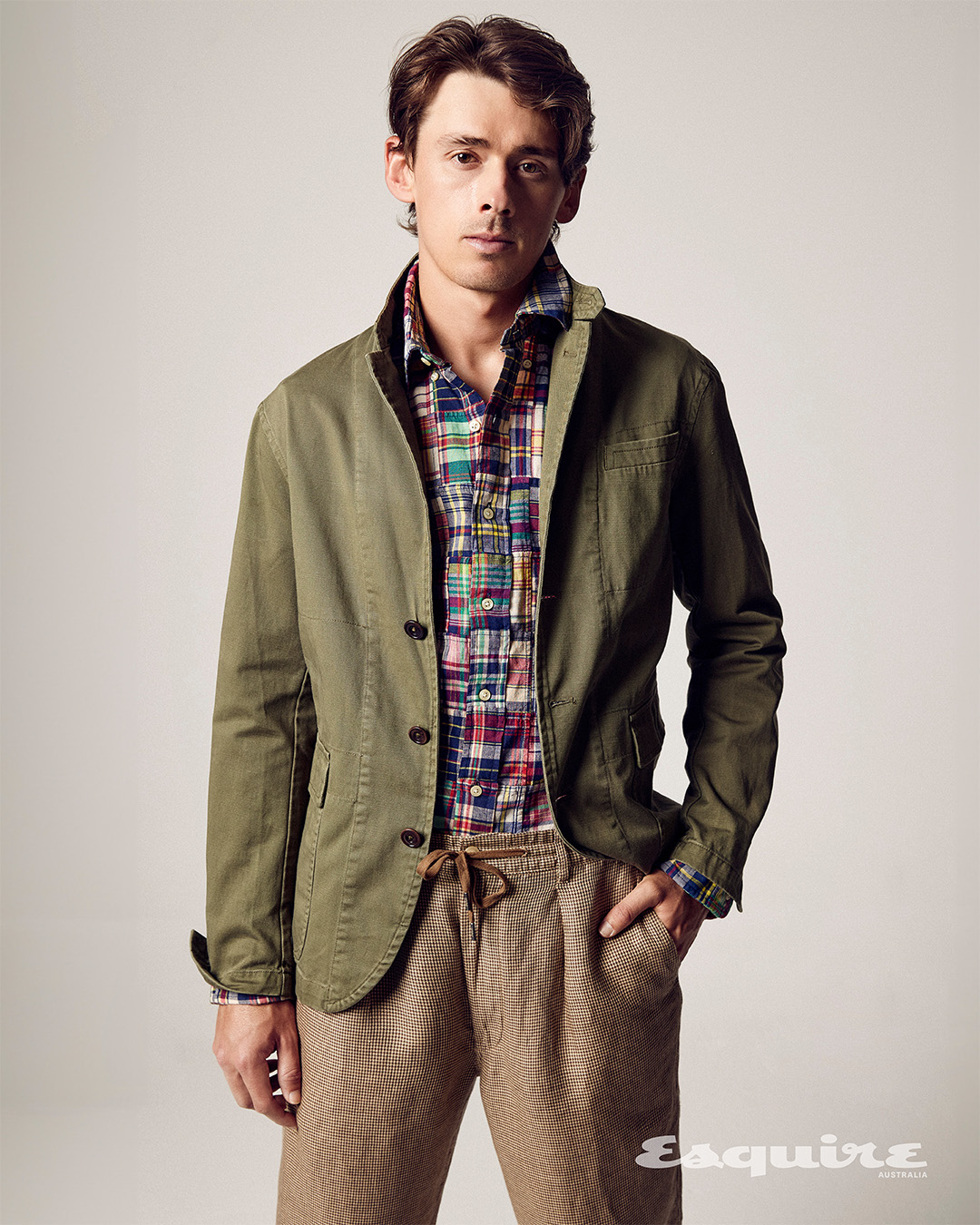 Portrait of a young adult man in a green jacket and colorful plaid shirt, posing in front of a neutral studio background with a magazine logo.