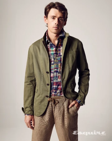 Portrait of a young adult man in a green jacket and colorful plaid shirt, posing in front of a neutral studio background with a magazine logo.