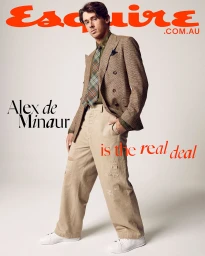 Alex de Minaur wearing a double-breasted brown blazer, patterned tie, and beige trousers against a white seamless background