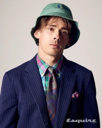 Alex De Minaur wearing a blue pinstripe suit, plaid shirt, paisley tie, and green bucket hat against a plain background.