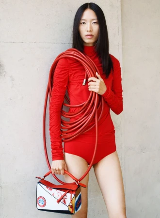 A young Asian woman in a red bodysuit carries a red hose and patterned purse against a neutral wall, showcasing minimalist fashion.