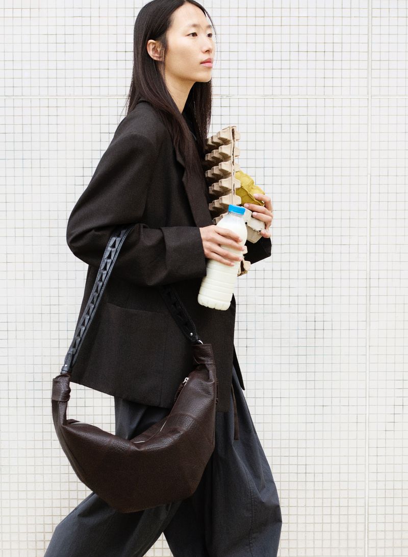 An Asian woman walks with a large brown bag, carton of eggs, and milk, wearing a black blazer and grey pants against a white tile background.