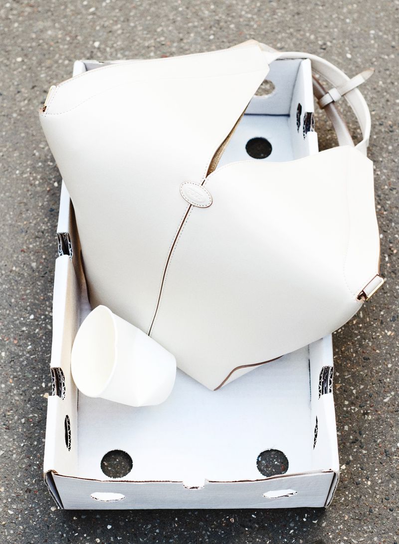 White leather handbag and cup inside a white cardboard box on a gray concrete surface, top-down view, product still life.