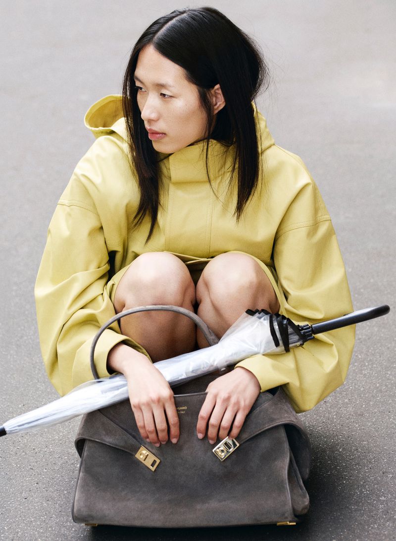 Asian woman crouches in a yellow hooded jacket, holding a grey satchel bag and a clear umbrella, set against a neutral grey background.