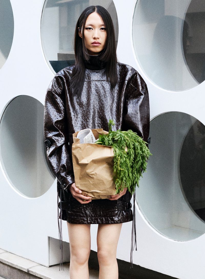 Asian model showcases a wet-look dark vinyl dress, holding a brown paper bag with greens, against a white geometric patterned wall.