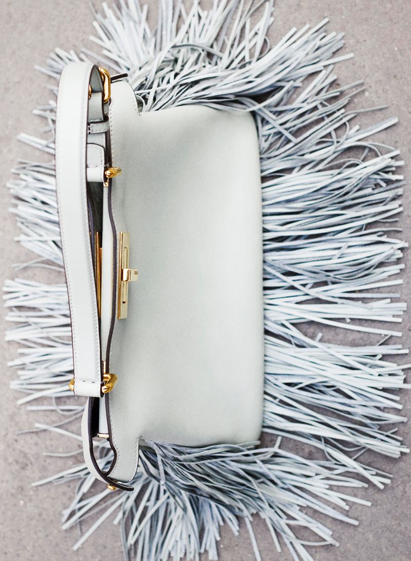 Close-up of a white leather crossbody bag with a gold clasp, surrounded by matching white fringe, set against a neutral background.