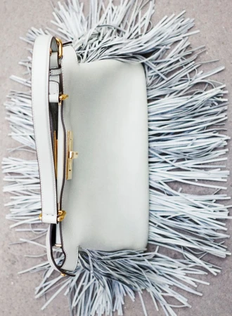Close-up of a white leather crossbody bag with a gold clasp, surrounded by matching white fringe, set against a neutral background.