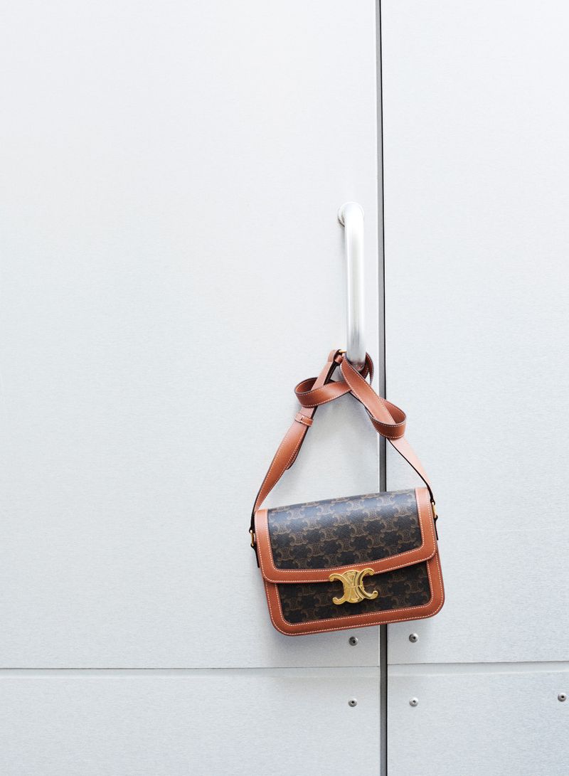 Luxury brown Celine handbag with the Triomphe logo hanging from a silver door handle against a plain white background in a studio.