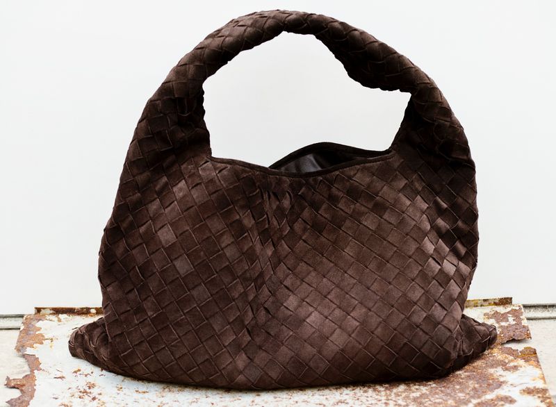 A brown woven leather handbag with a braided handle sits on a rusty metal surface against a white backdrop in a studio setting.