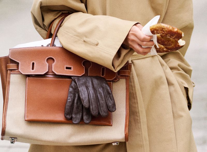 Tan trench coat with a tan and brown handbag with black leather gloves, and a person eating a pastry, neutral outdoor setting.