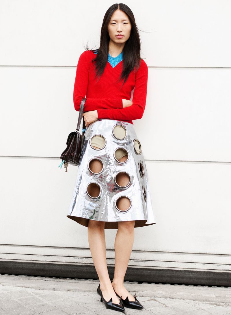 An Asian woman models a metallic skirt with cutouts, paired with a red sweater and black heels, set against a white wall backdrop.