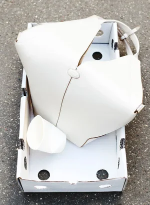An off-white leather tote bag and a white cup resting inside an open white cardboard tray on dark grey asphalt.