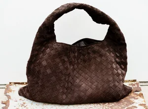 A woven dark brown suede hobo bag rests on a weathered rusty surface against a plain white background.