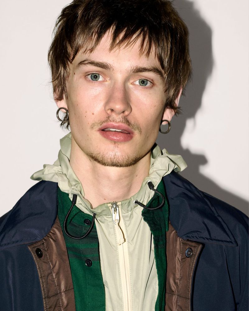 Close-up portrait of a young man wearing layered outerwear with hoop earrings against a white studio background.