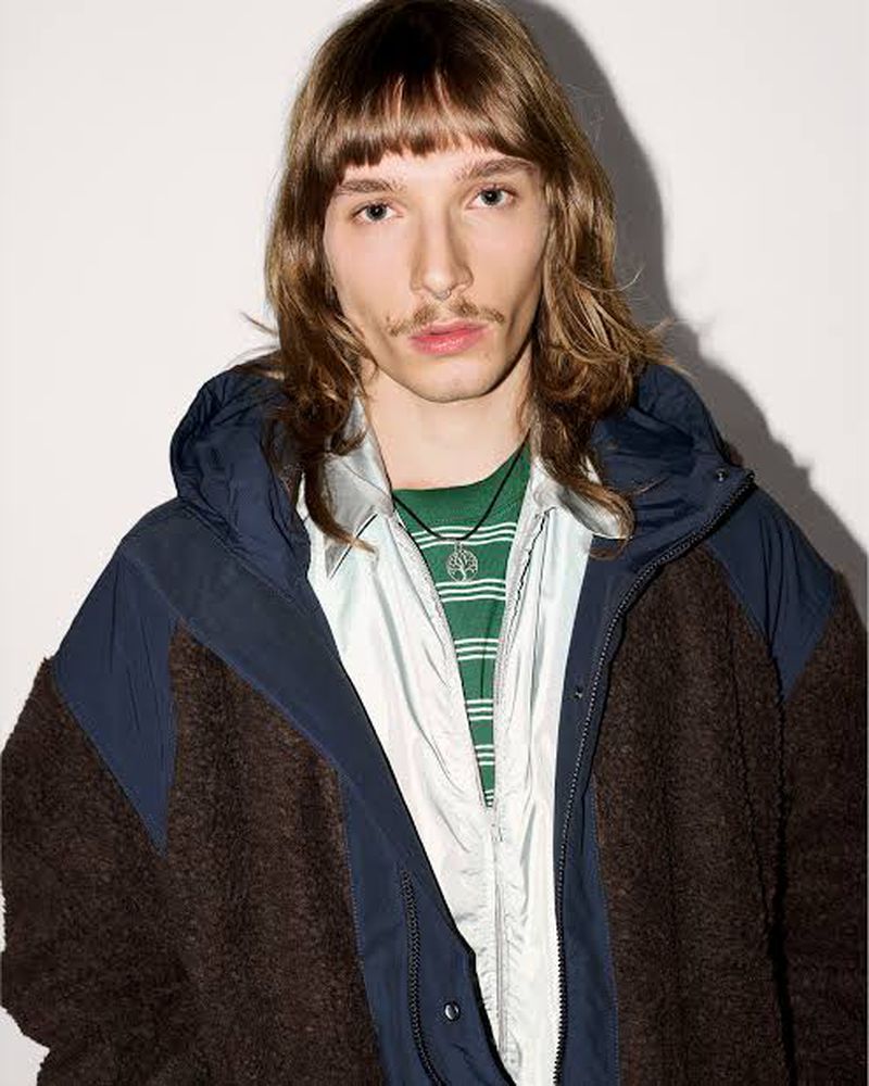Model with long hair and mustache wearing a layered Canada Goose fleece jacket over a striped green shirt against a white backdrop.