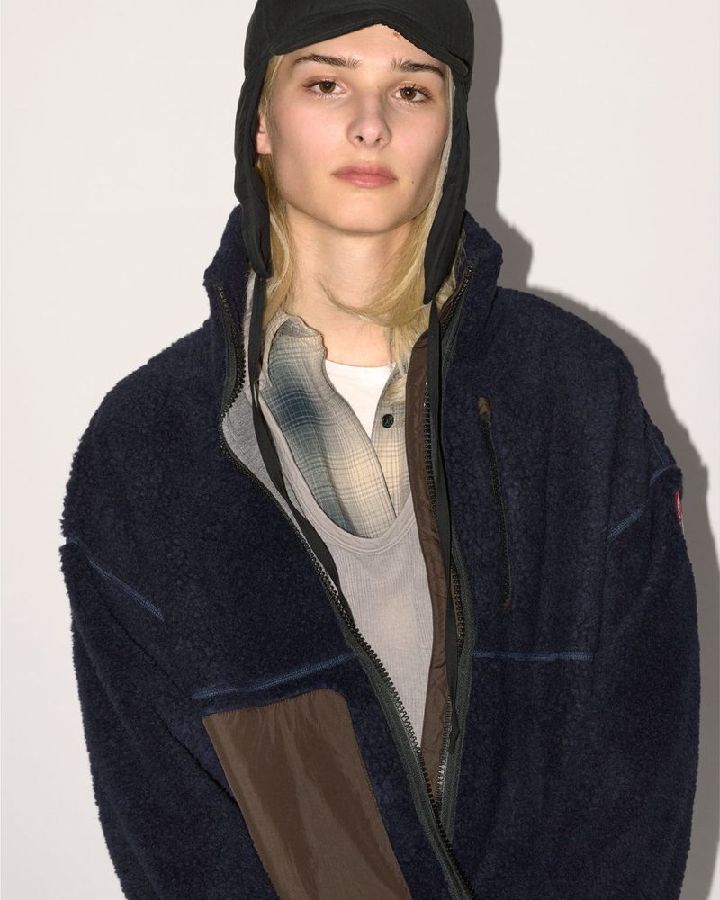 Model wearing a dark navy fleece zip jacket and black ear-flap cap against a light background for Canada Goose.