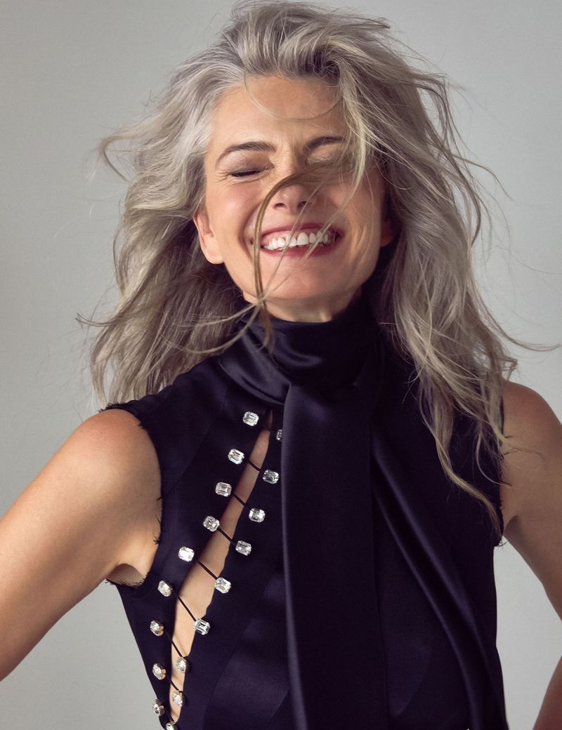 Close-up of a mature woman with long gray hair, smiling with eyes closed, wearing a black sleeveless top with rhinestone embellishments.