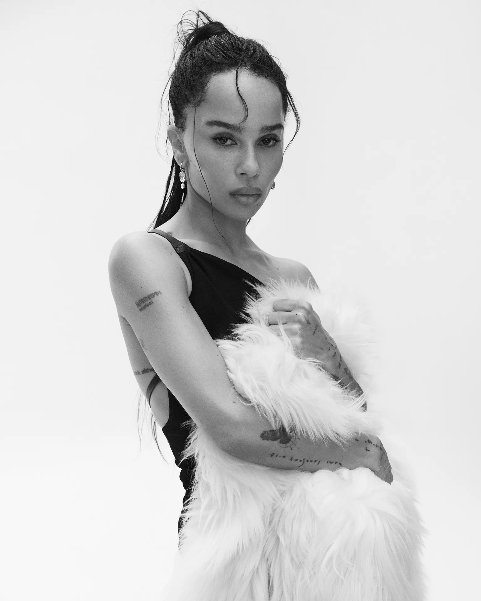 Zoë Kravitz holding a textured white faux fur wrap while wearing a black one-shoulder top against a white studio background.