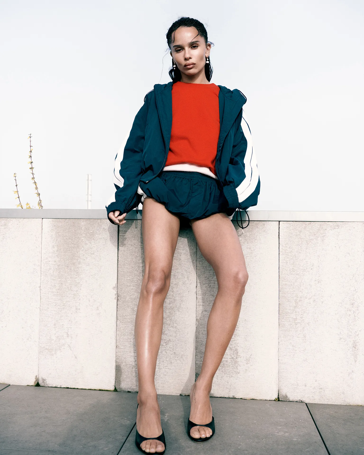 Zoë Kravitz leaning against a concrete wall wearing a teal windbreaker jacket, red top, shorts, and black heels.