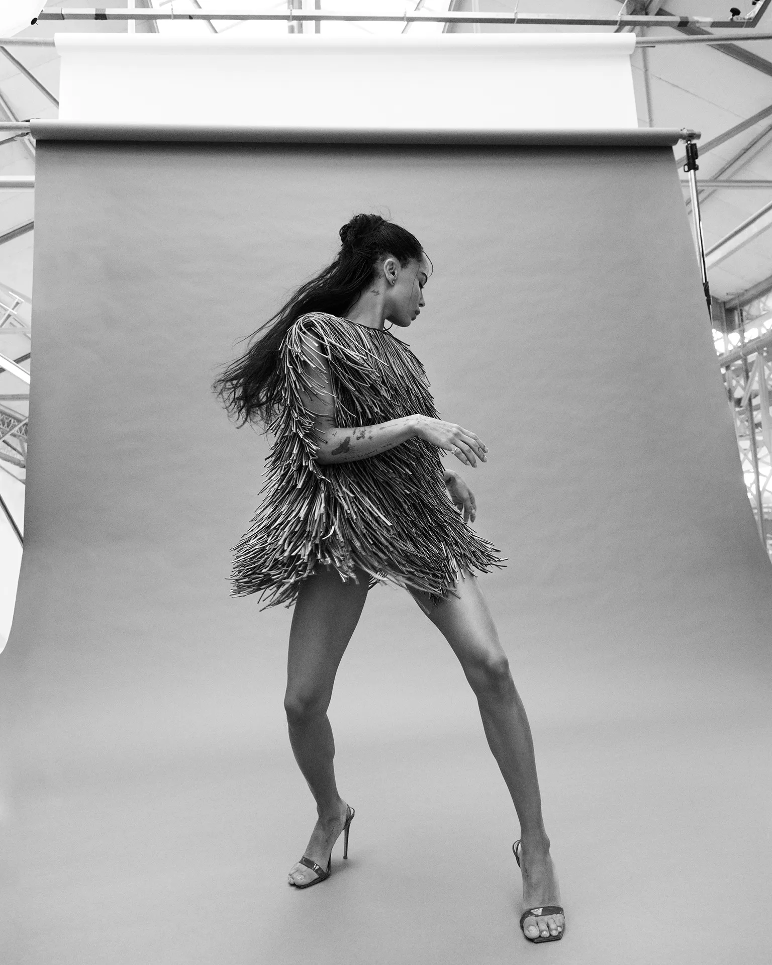 Zoe Kravitz in a fringed dress and heels posing against a grey studio backdrop captured by Mikael Jansson for British Vogue.