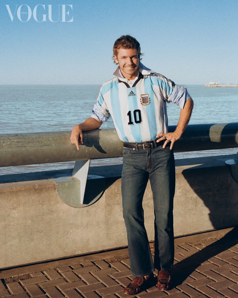 Man in a number 10 Argentina soccer jersey and denim jeans smiling by a waterfront railing for Vogue Latin America.