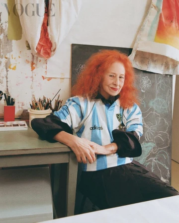 A woman with voluminous bright orange hair wearing a light blue and white striped Argentina soccer jersey seated at an art studio desk.