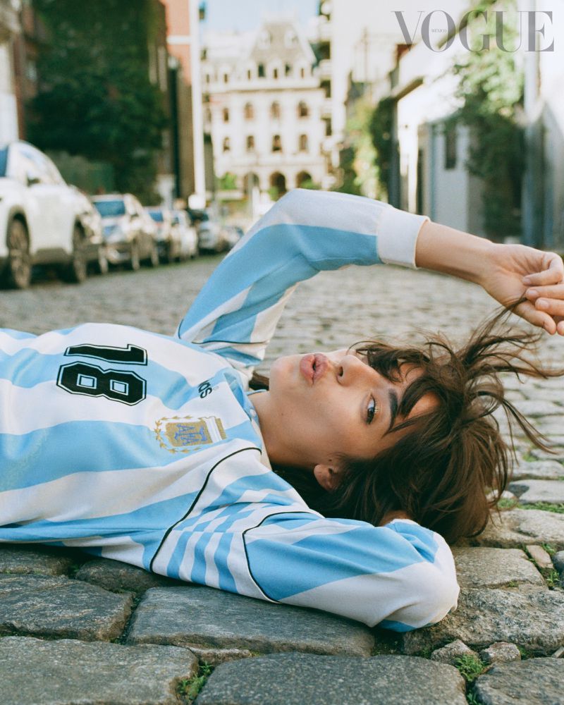 A woman lying on a sunlit cobblestone street wearing a blue and white striped Argentina football jersey while tugging her hair.