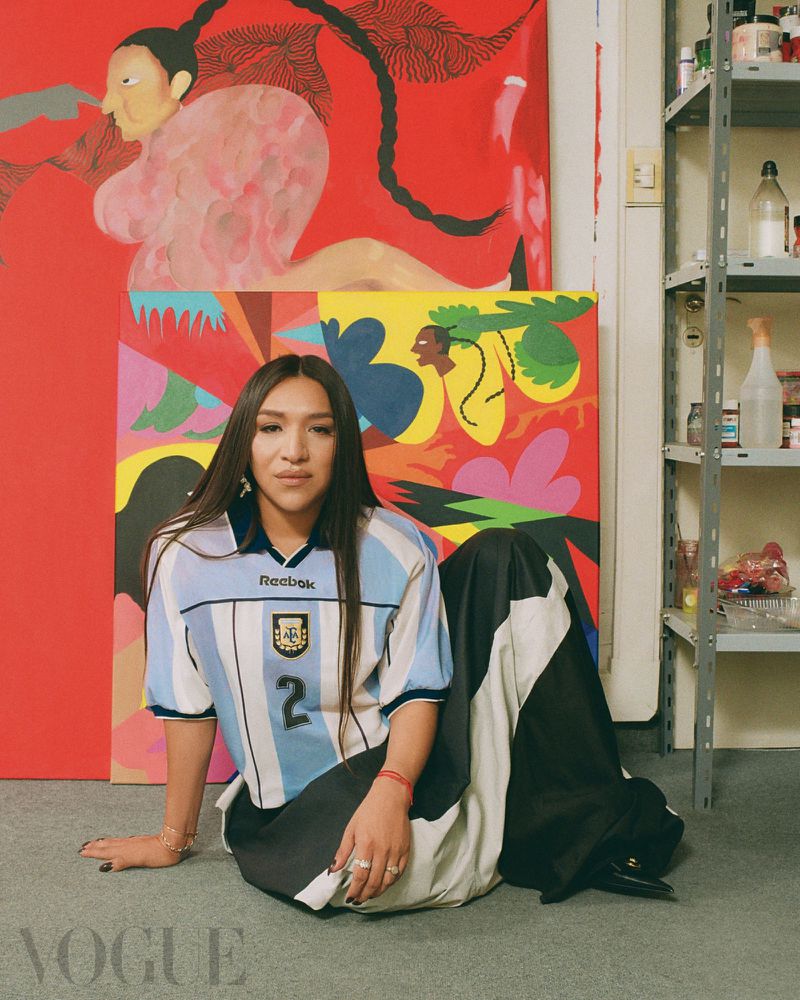 Person in a vintage Argentina national football team jersey and wide skirt seated in front of vibrant artwork.