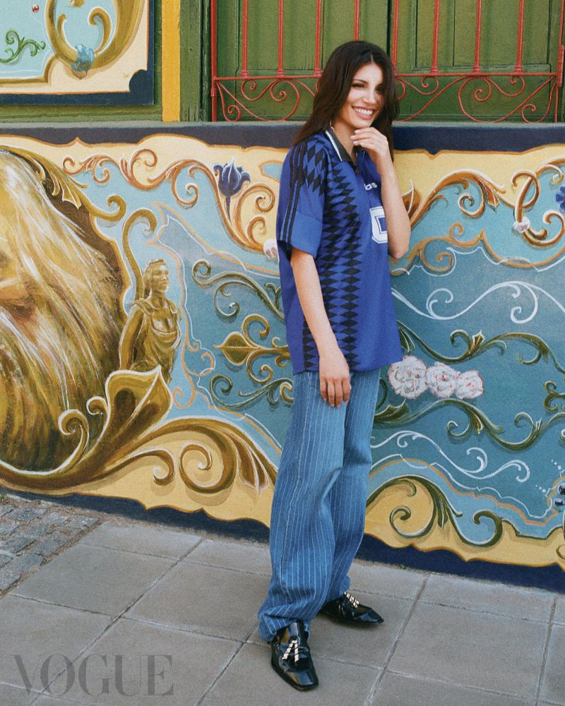 A smiling woman posing in a blue patterned football jersey and wide-leg denim jeans against a colourful painted street mural.