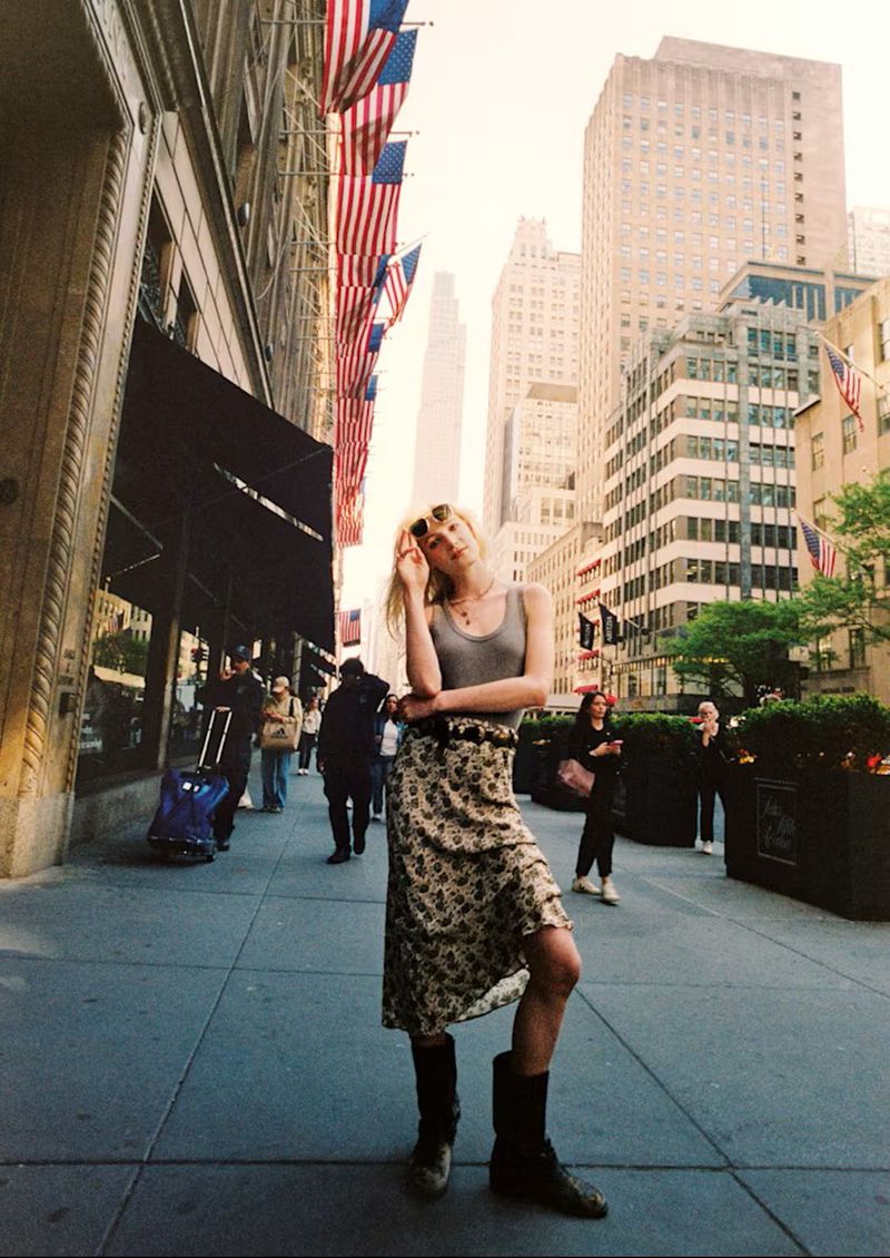Model Summer Dirx posing on a city sidewalk wearing a grey tank top, floral midi skirt, and black boots beneath hanging American flags.