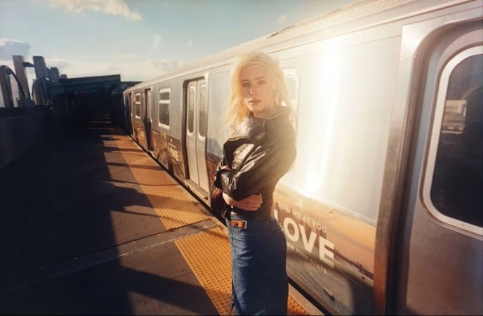 Blonde model Summer Dirx wearing a black leather jacket and denim skirt on an outdoor train platform in golden sunlight