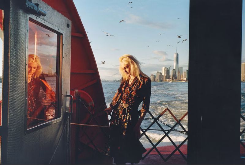 Model Summer Dirx standing on a ferry deck in a dark patterned dress with the Manhattan skyline across the water in golden sunlight.
