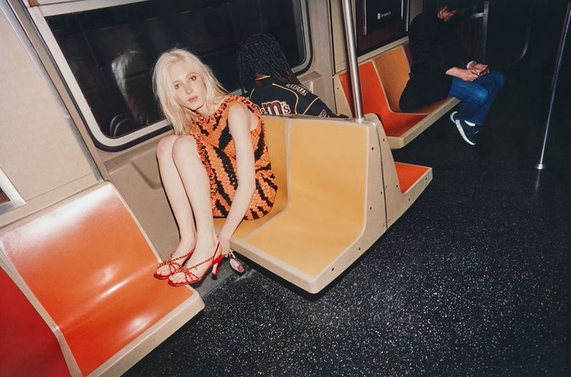 Model with blonde hair wearing an orange and black patterned dress sitting on a subway car seat