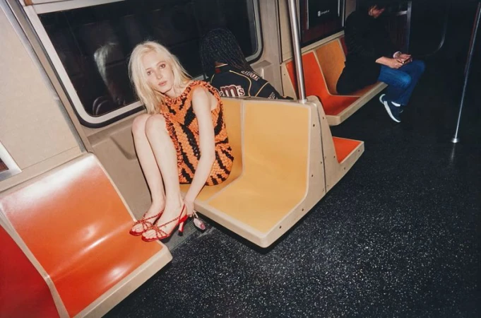 Model with blonde hair wearing an orange and black patterned dress sitting on a subway car seat