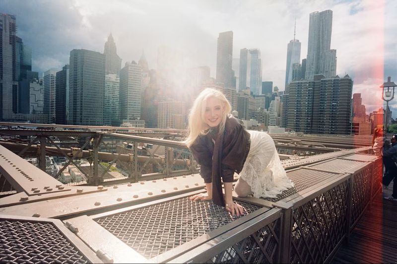 Blonde woman crouching on the Brooklyn Bridge railing in a brown jacket and white lace dress with the Manhattan skyline behind her.