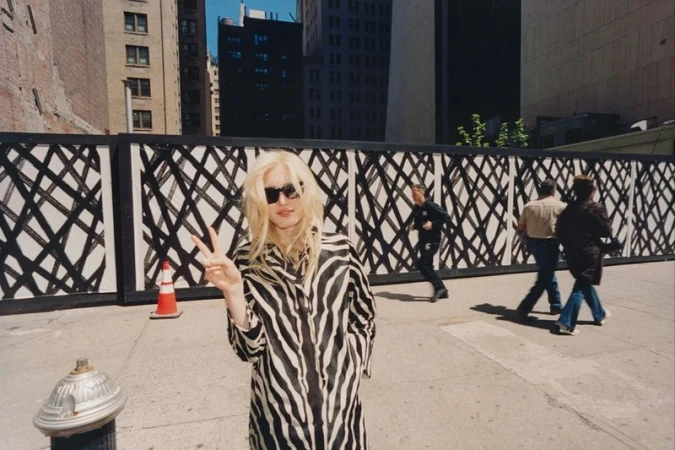 Summer Dirx in a zebra-print coat and sunglasses flashing a peace sign on a city sidewalk in front of a patterned construction wall.