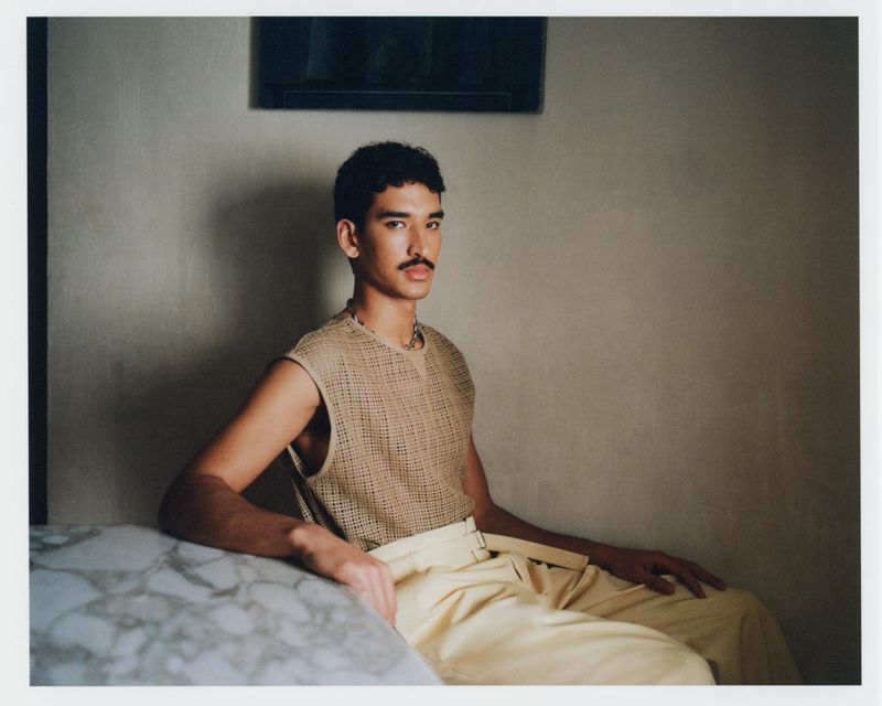 Man with mustache wearing a sleeveless beige knit top and high-waisted trousers seated beside a marble table.