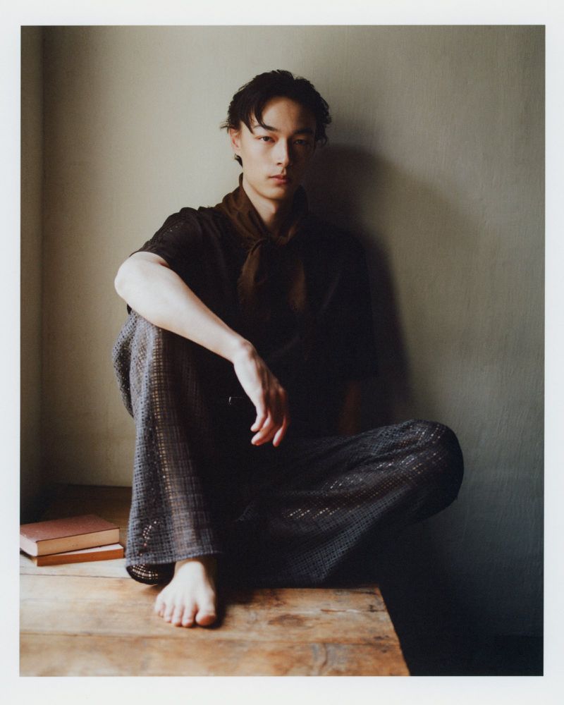 Barefoot male model seated on a wooden floor in dark textured trousers and a neck scarf against a neutral interior wall.