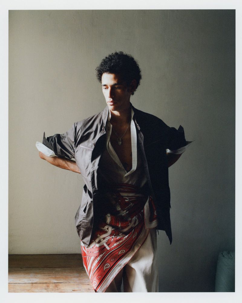 A man standing with hands behind his back wearing a dark jacket, cream trousers, and a patterned red silk scarf tied at his waist.