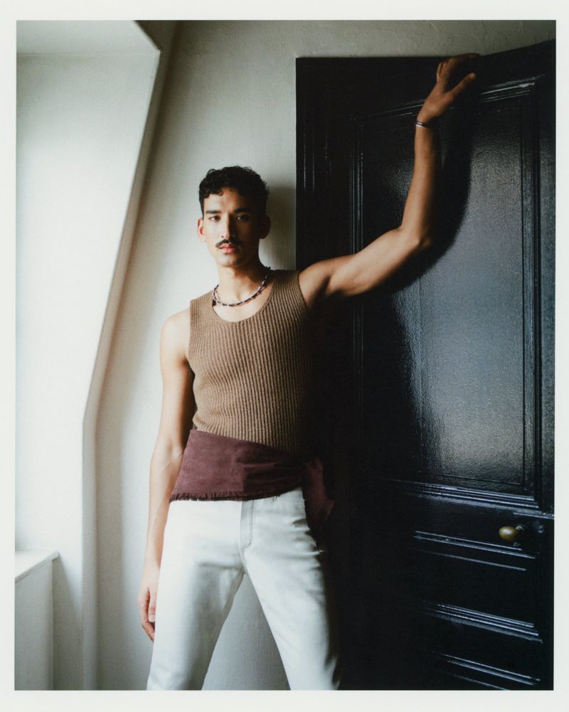 A man in a brown ribbed sleeveless tank top and white trousers posing beside a dark painted interior door.