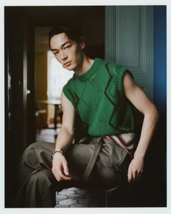 Model seated wearing a bright green diamond-knit sleeveless vest and pleated taupe trousers by an open interior doorway.