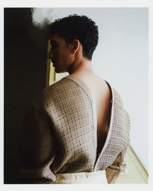 Man viewed from behind looking sideways in an open-back beige textured woven top lit by window light.
