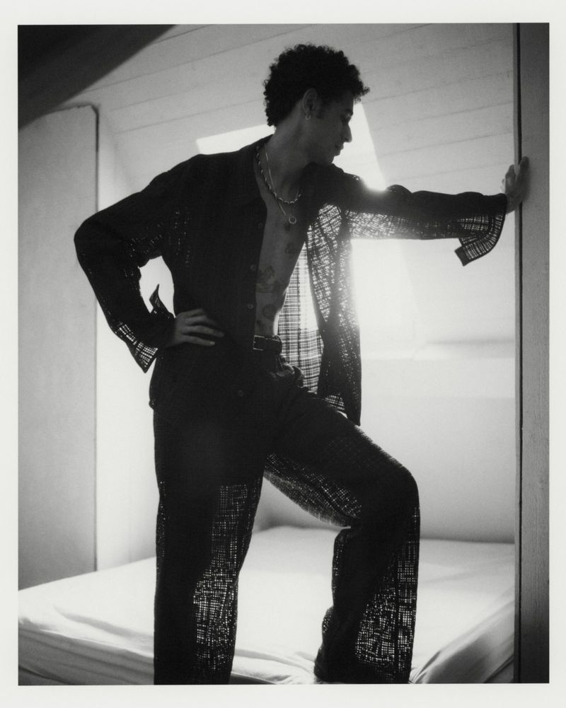 Model posing on a bed wearing an open sheer black patterned shirt and trousers in a backlit black and white fashion editorial.