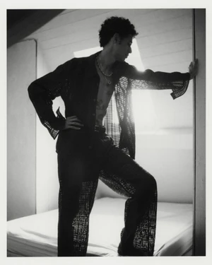 Model posing on a bed wearing an open sheer black patterned shirt and trousers in a backlit black and white fashion editorial.