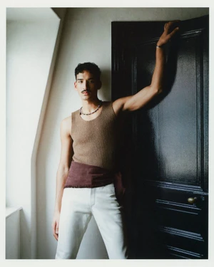 A man in a brown ribbed sleeveless tank top and white trousers posing beside a dark painted interior door.
